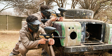 Two men crouching with paintball guns against an old jeep